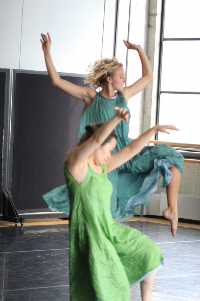 In a lime green dress, one dancer lifts both arms up. Behind her, in a forest-green dress, another dancer lifts one leg, which is bent at the knee, and both arms.